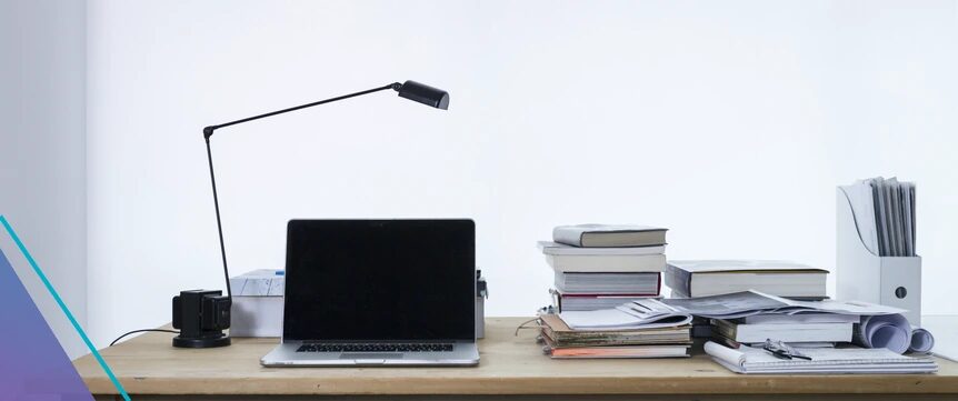 A bright study desk with an open laptop, a black desk lamp and stacks of books and papers against a plain white wall